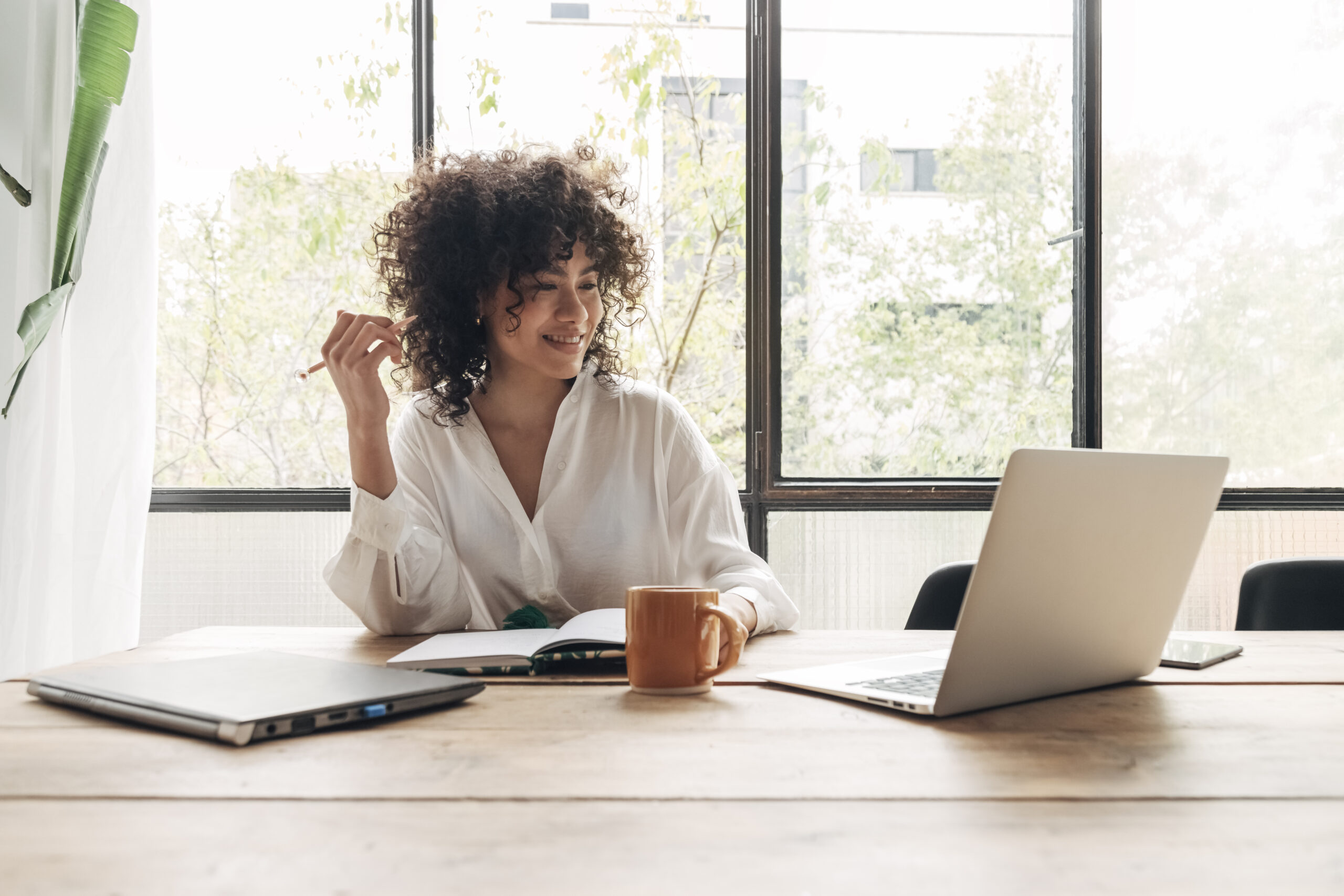 A woman is smiling and sitting at her desk with her laptop open while writing in her notebook.