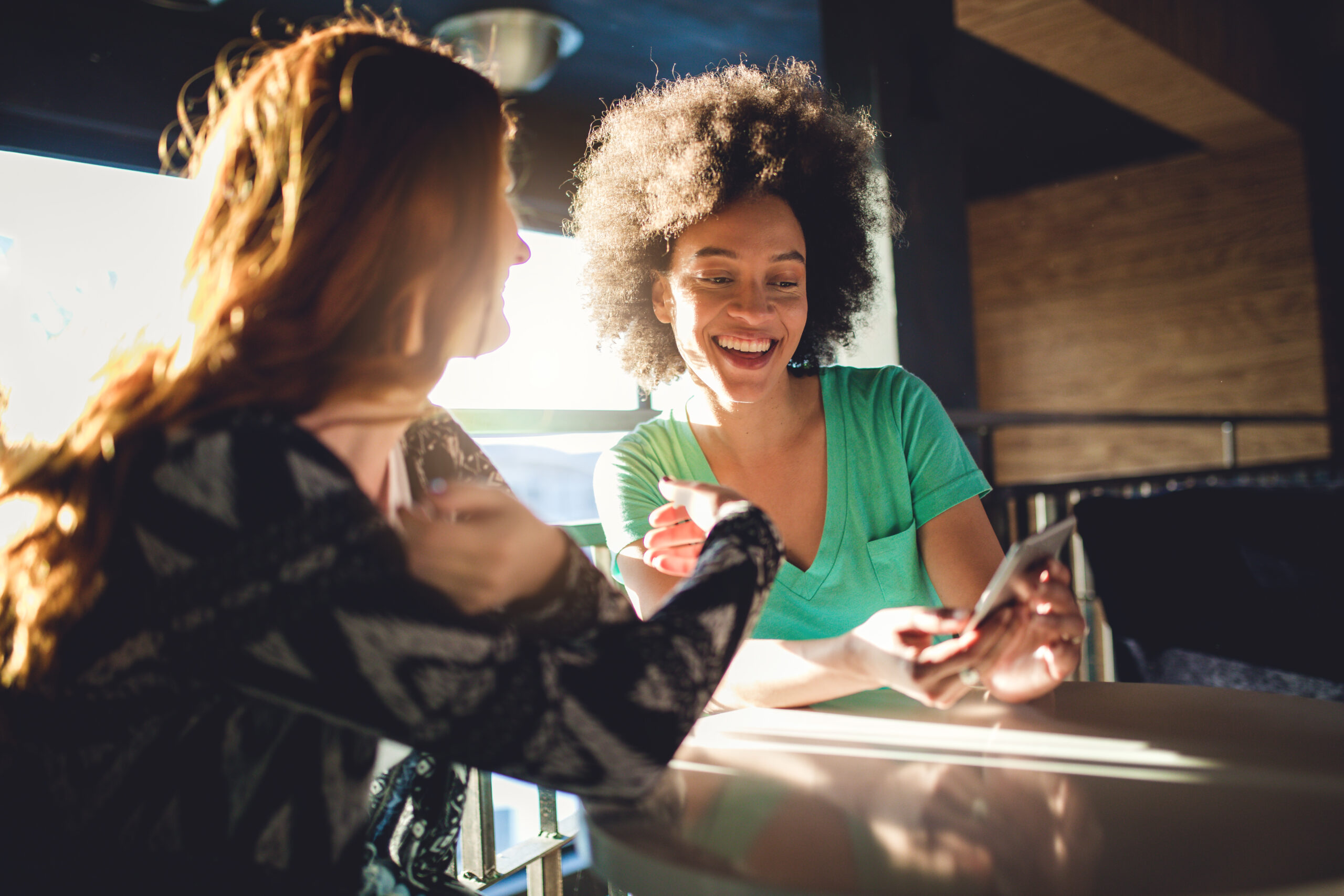 Two female friends sitting in a cafe and looking at their mobile phone while smiling.